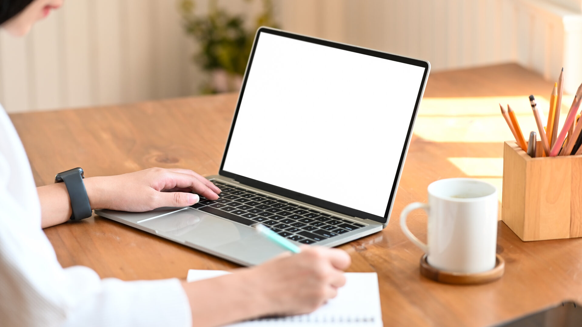 Cropped shot of young designer woman writing on notebook while s Cropped shot of young designer woman writing on notebook while she sitting and using white blank screen laptop at the modern wooden table. Women lifestyle concept.